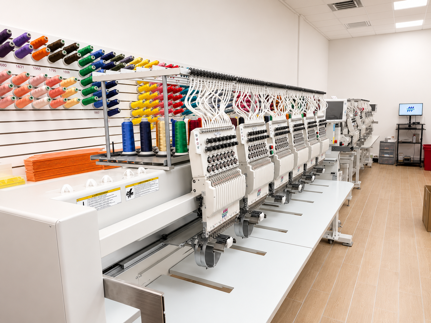 A long row of white industrial embroidery machines on a white table, each mounted with multiple colorful thread spools, photographed in a bright clean studio or workshop with wood flooring.
