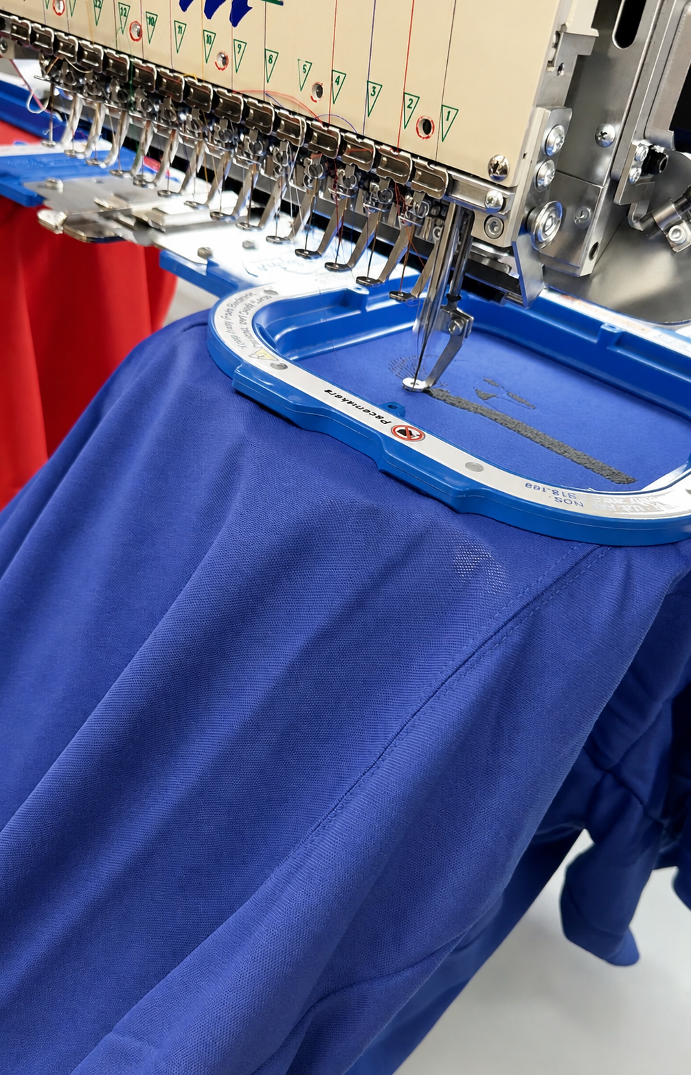 Close-up of an industrial multi-head embroidery machine stitching a royal blue fabric garment inside a blue hoop.