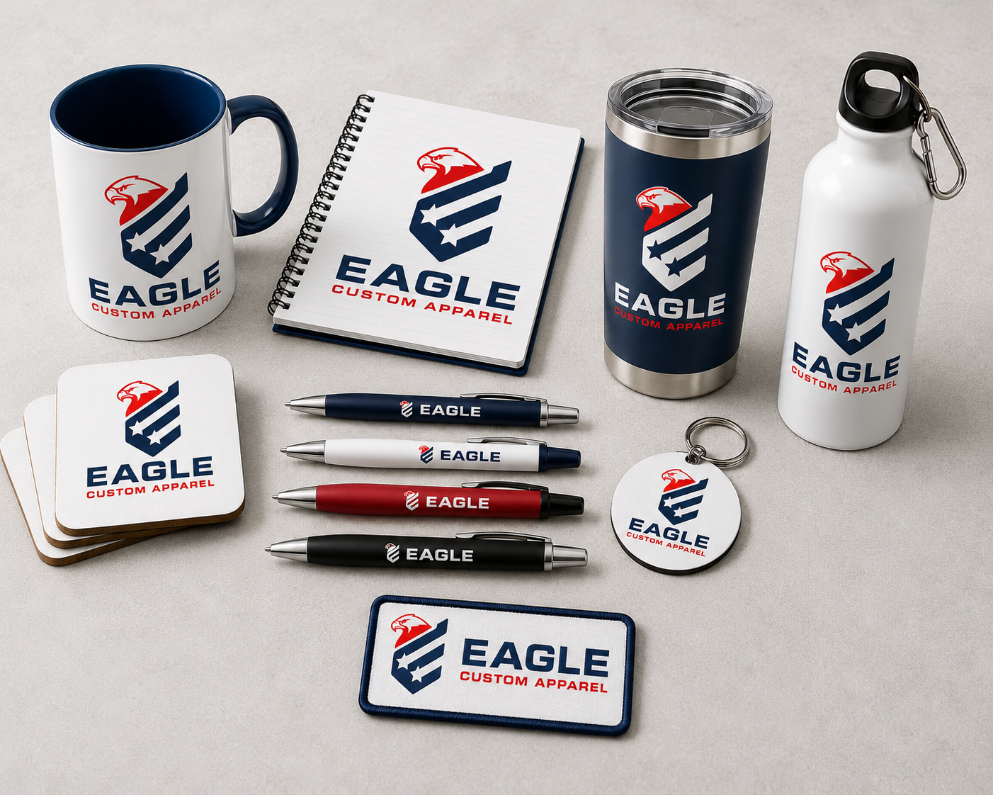 A coordinated set of branded Eagle Custom Apparel merchandise arranged on a light neutral surface, including a mug, notebook, insulated tumbler, water bottle, coasters, pens, a round keychain, and a rectangular patch or sticker. The items all show the same eagle-and-flag style logo with the text EAGLE and EAGLE CUSTOM APPAREL.