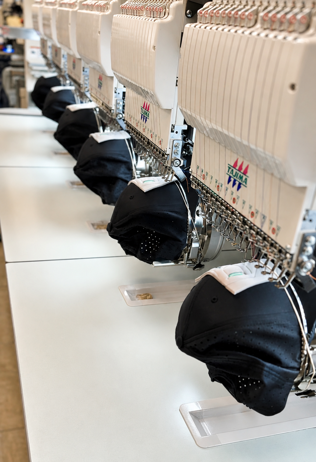 A close-up view of multiple industrial embroidery machines lined up on a white table, each holding a black fabric cap or bandana under the needle area.