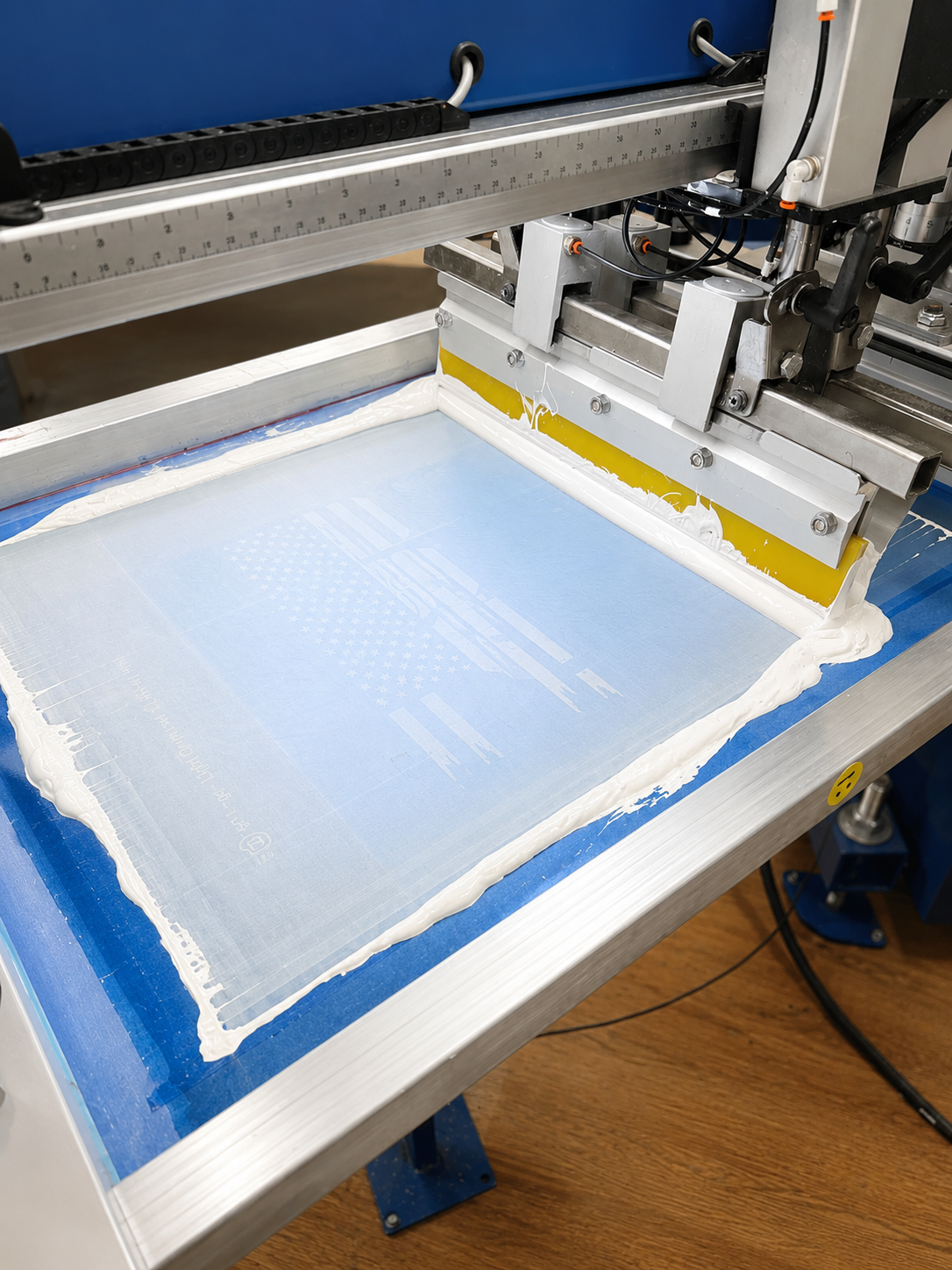 A close-up view of a screen printing machine with a white mesh screen and blue tape, positioned over a metal printing bed on a workshop floor.