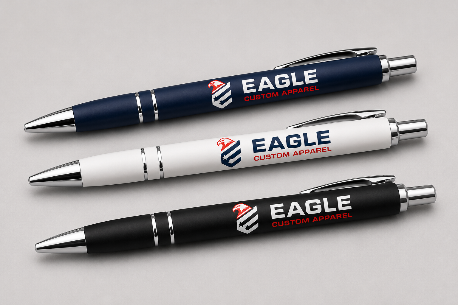 Three custom-printed ballpoint pens in navy blue, white, and black, each featuring the Eagle Custom Apparel logo and text, arranged diagonally on a light gray background.